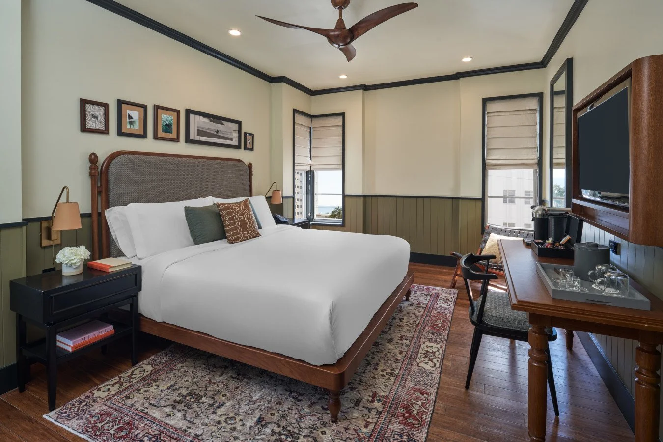 A hotel room with a large bed, side tables, Wall art, multiple windows, a wall-mounted TV, a desk with a coffee maker, and a ceiling fan.