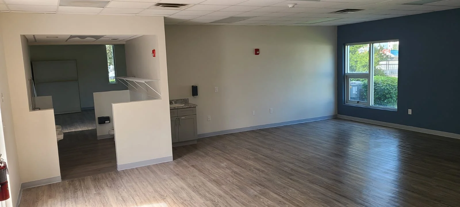 An empty room with large windows, wood flooring, and beige and dark blue walls, with a small kitchenette area on the left.