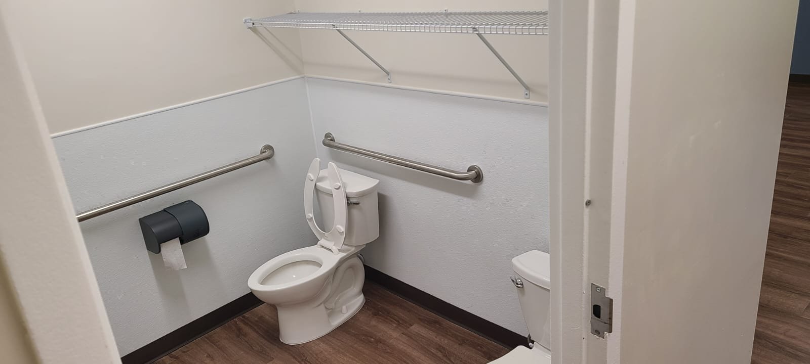 Accessible bathroom with white toilet, metal handrails, a black toilet paper holder, wall-mounted shelf, and wood-style flooring.