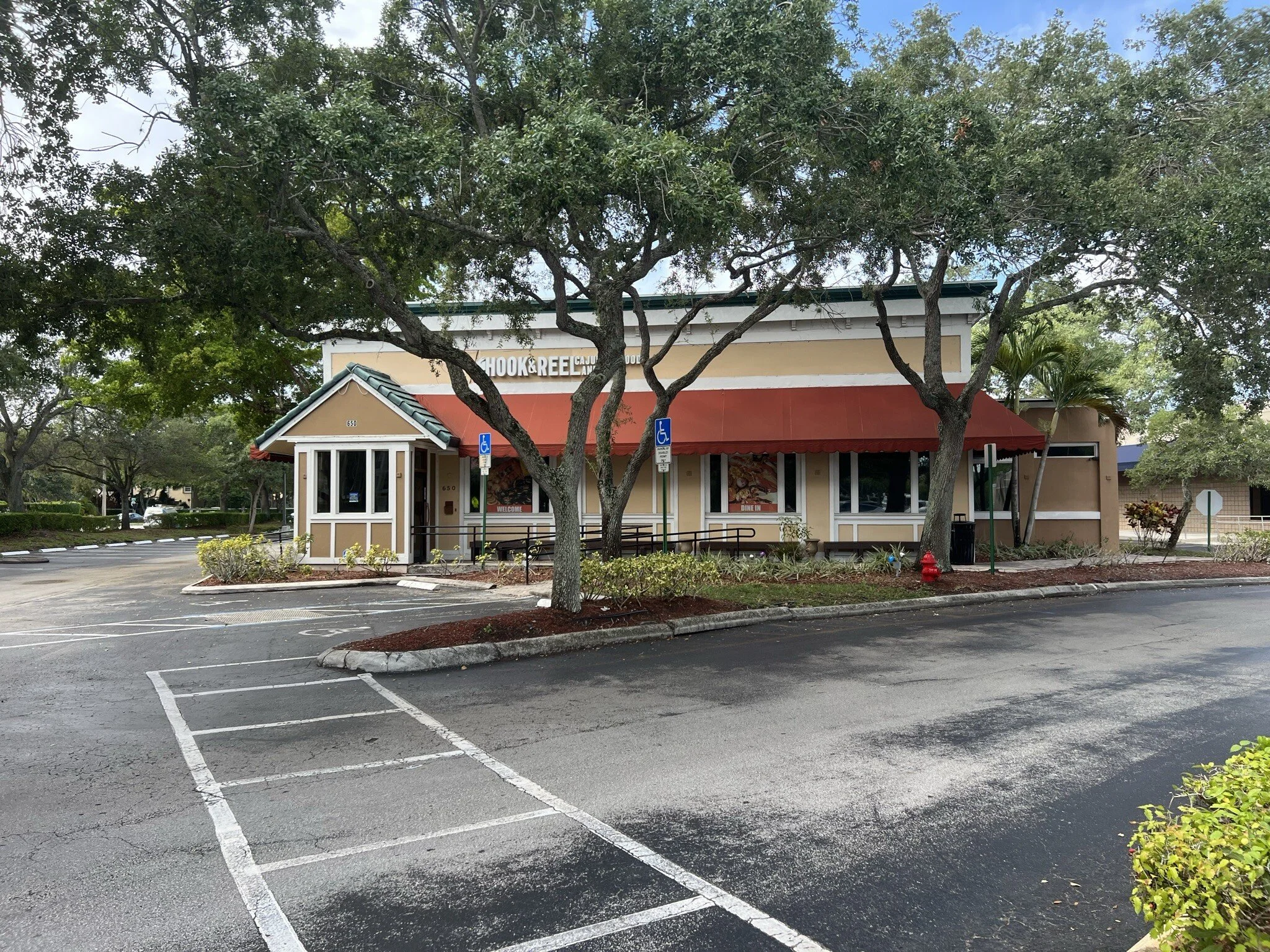 Empty parking lot in front of a California-style restaurant with a red awning, beige walls, and white trim. The restaurant has a sign that reads 'Hook & Reel Cajun Seafood,' and there are trees and plants surrounding the building.