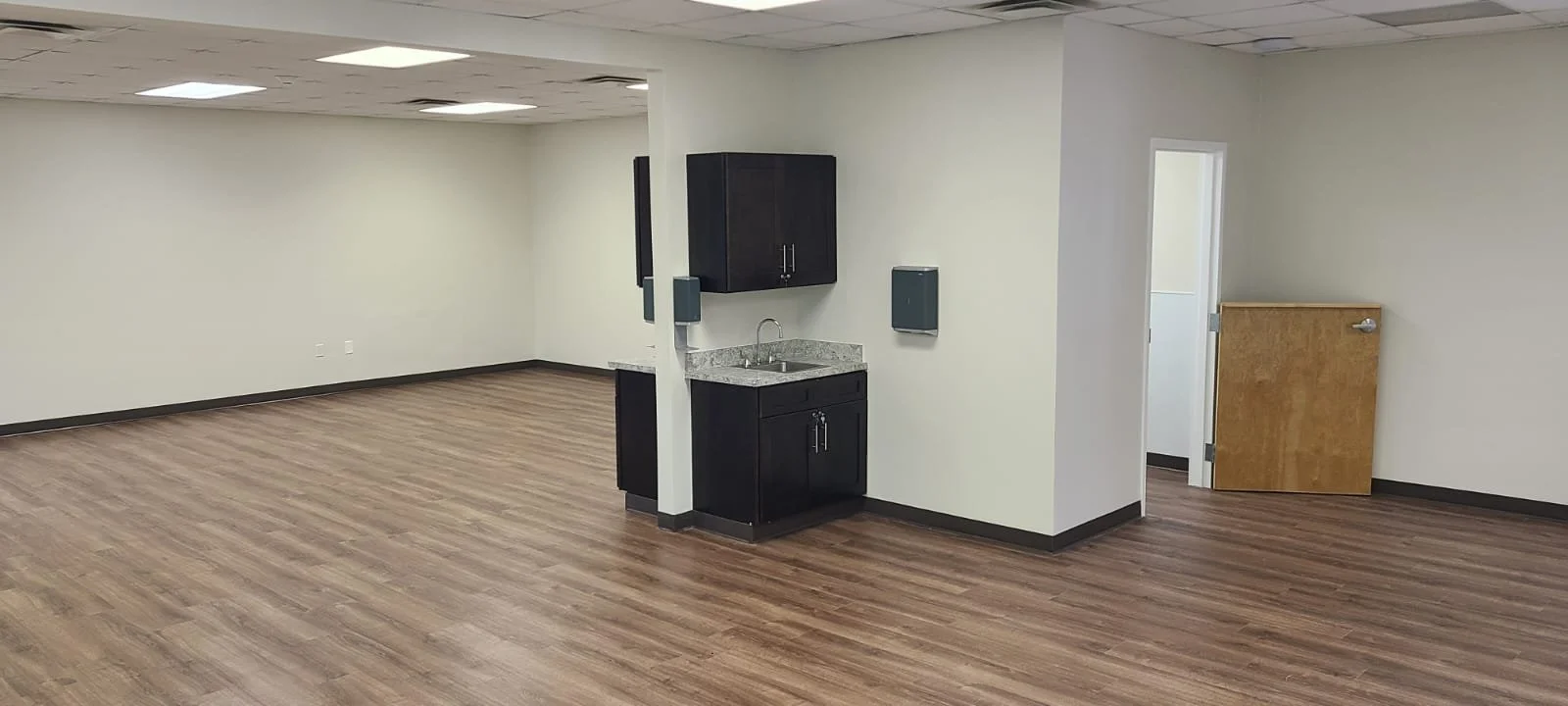 Empty room with wooden flooring, plain white walls, ceiling lights, small kitchenette with dark cabinets, granite countertop, sink, and wall-mounted speakers.