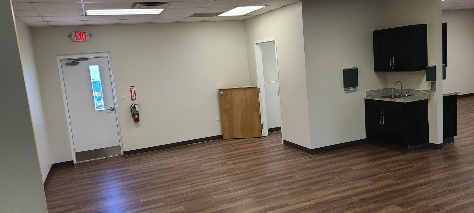 Empty room with wooden floor, a closed door with an emergency exit sign, and a small kitchenette with dark cabinets, granite countertop, and a sink.