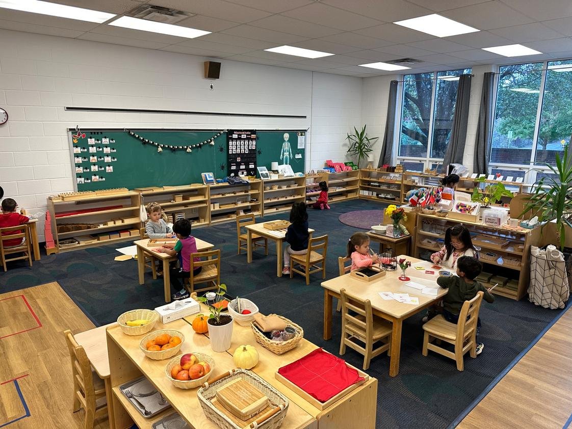 A classroom with children engaging in various activities at tables and shelves, decorated with fall produce and plants, featuring large windows letting in natural light.