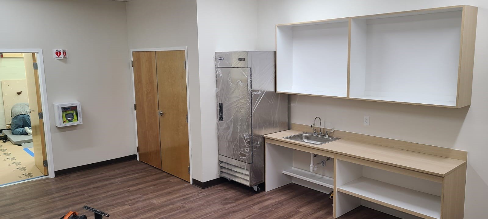 Empty kitchenette with wooden countertop, white cabinets, stainless steel refrigerator wrapped in protective plastic, sink, and open shelving in a room under construction.