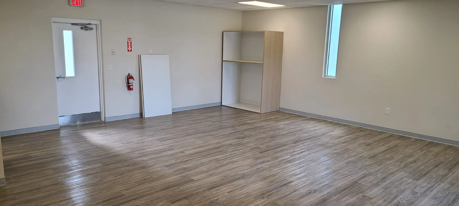 An empty room with beige walls, light wood flooring, a door with a small window, a fire extinguisher, a whiteboard, a wooden shelving unit, and a narrow window.