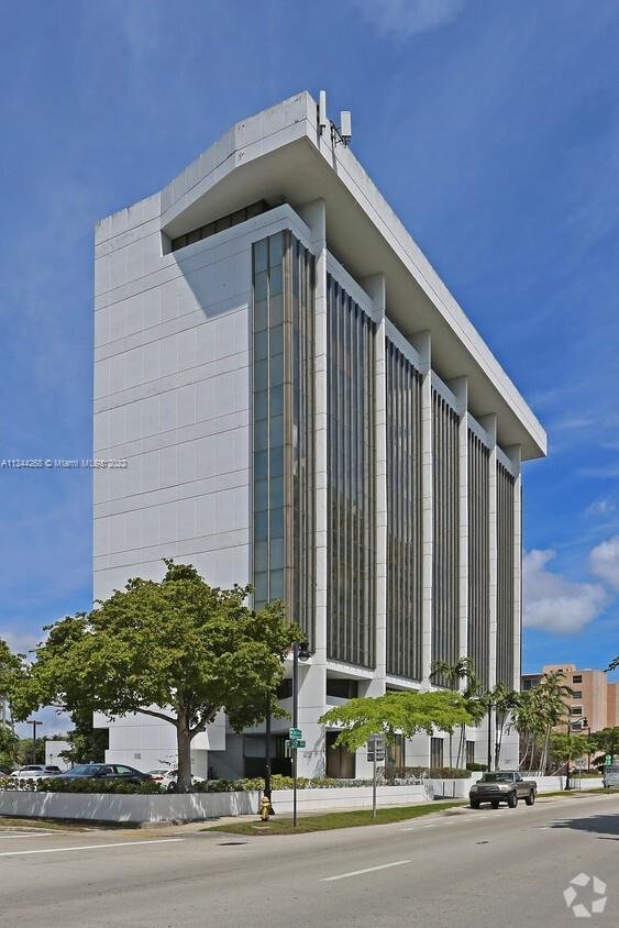 Modern high-rise building with vertical glass windows and white facade, surrounded by trees and cars on a city street under a blue sky.