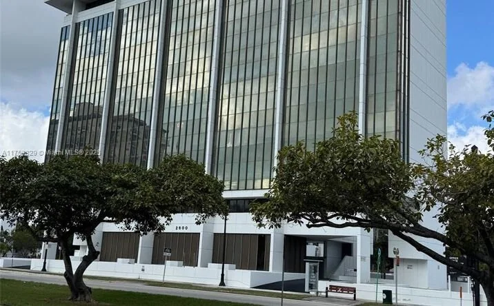 Tall modern glass office building with trees and a sidewalk in front.