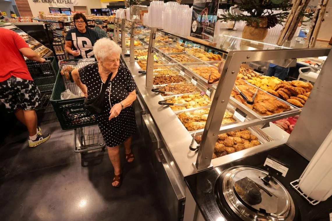 Elderly woman in a black dress with white polka dots shopping at a bakery counter in a grocery store. The bakery display case has various baked goods including cookies, bread, and pastries. Other shoppers are visible in the background.