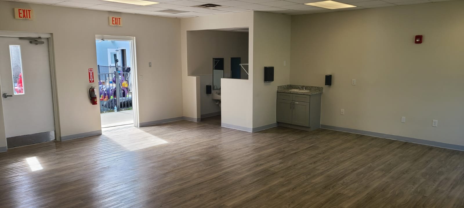 Empty room with wood laminate flooring, white walls, two exit signs, a fire extinguisher, an open door leading outside, and a corner with a small sink and countertop.