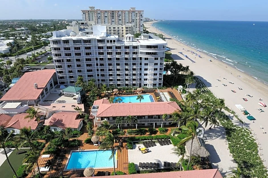 Aerial view of a beachfront hotel with swimming pools, palm trees, and a sandy beach along the ocean.