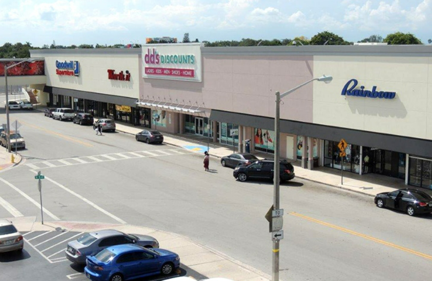 A shopping mall exterior with various store signs, including Goodwill, Murphy's, dd's Discounts, Rainbow, and picnic-pavilion style seating outside.