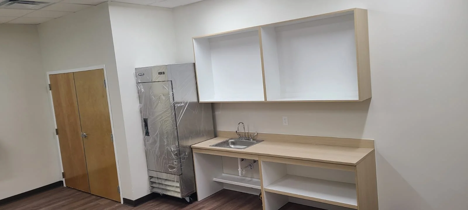 Empty kitchenette with wood cabinets, a stainless steel refrigerator wrapped in protective plastic, and a small sink, in an unfurnished room with white walls and wood flooring.