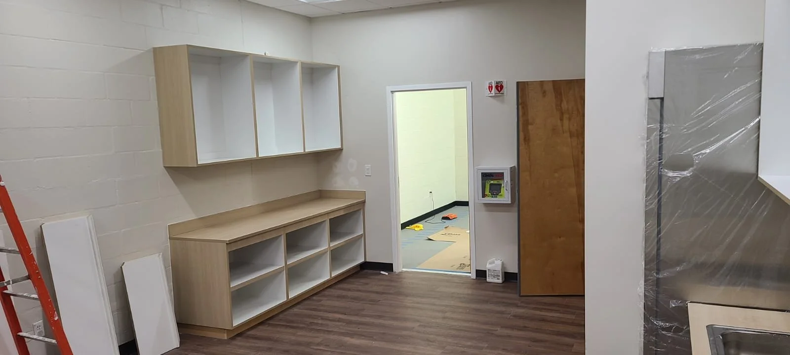 Interior of a room under construction with wooden shelves, an open doorway leading to a room with flooring, a ladder on the left, and construction tools on the floor.