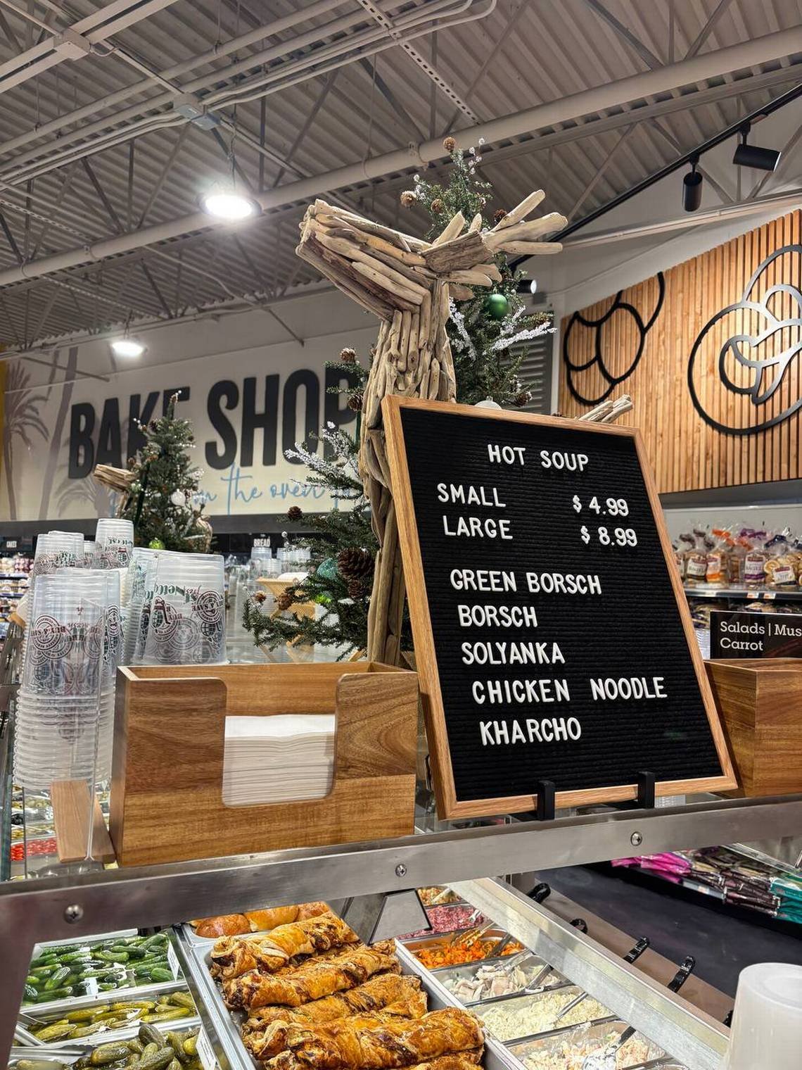A black menu board with white text listing hot soup options and prices, decorated with a wooden reindeer sculpture made of driftwood and a small Christmas tree in the background at a bakery shop.