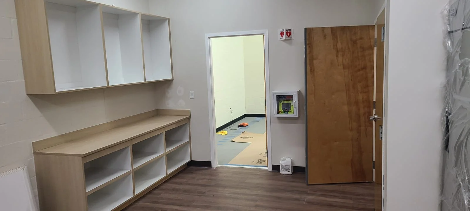 Room with white and wood-colored shelves, a doorway leading to another room with a yellow wall and a black and gray floor, a wooden door leaning against the wall, and a box of cleaning supplies on the floor.