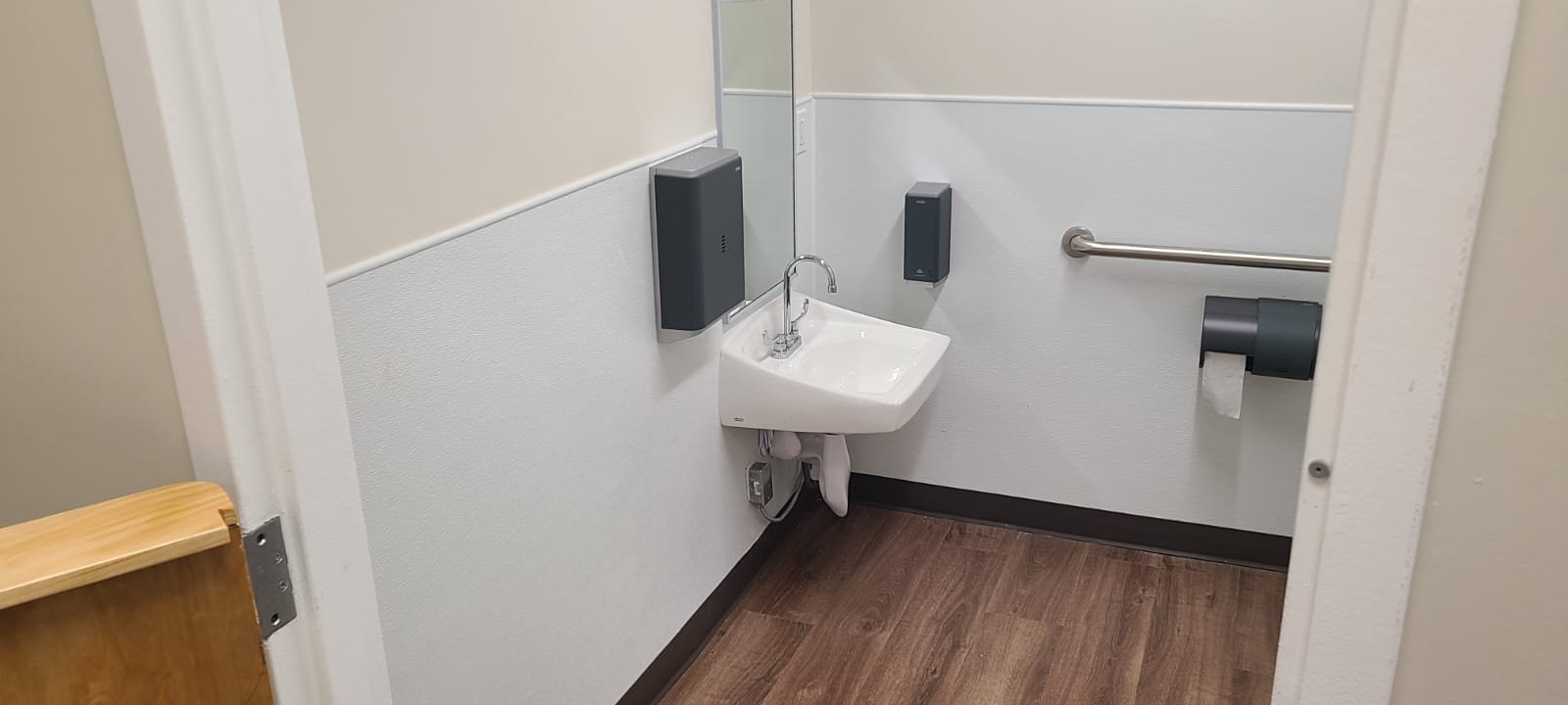 Small bathroom with a wall-mounted sink, a mirror, a soap dispenser, a paper towel dispenser, a hand dryer, and a wall-mounted grab bar on the right side.
