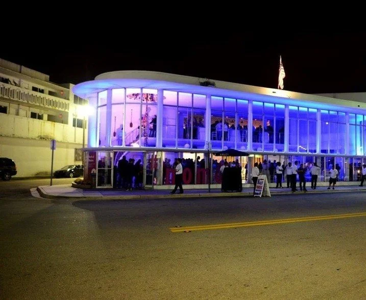 Glass building with blue and purple lighting at night, filled with people inside and outside.
