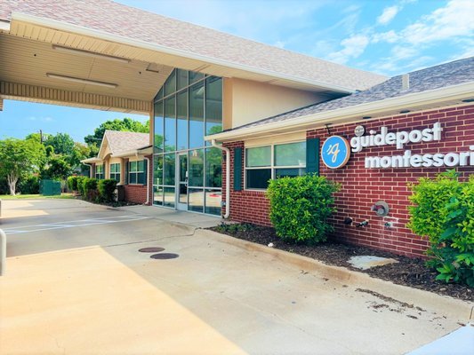 The entrance of Guidepost Montessori with a brick building, large glass windows, and a sign displaying the Guidepost Montessori logo. A covered walkway and sunny weather with a blue sky are visible.