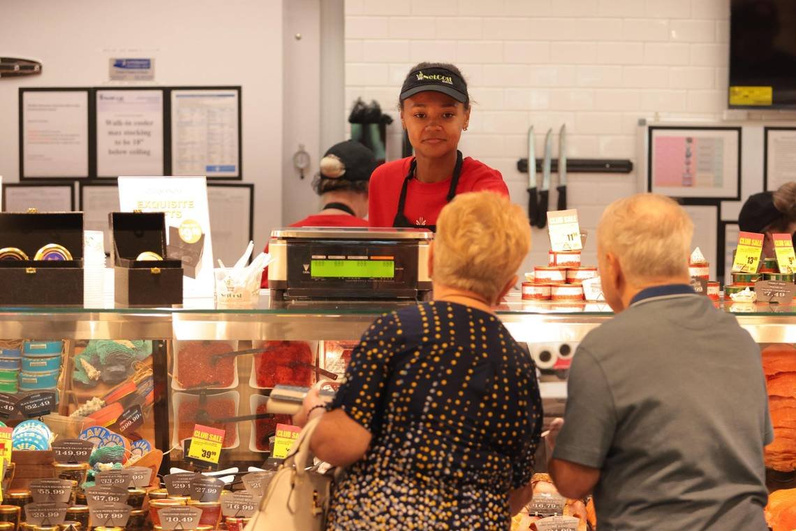 A seafood counter at a store with two elderly customers ordering, an Asian female employee behind the counter looks on, and various seafood products and price tags are visible.