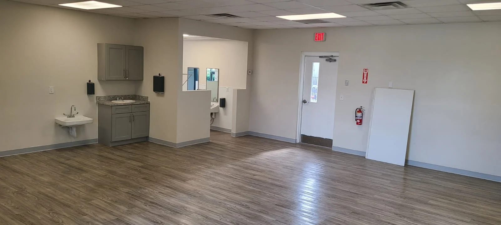 Empty room with wood flooring, small sink area, wall-mounted cabinets, and a handicapped accessible sink. Exit door, fire extinguisher, and whiteboard on the wall.