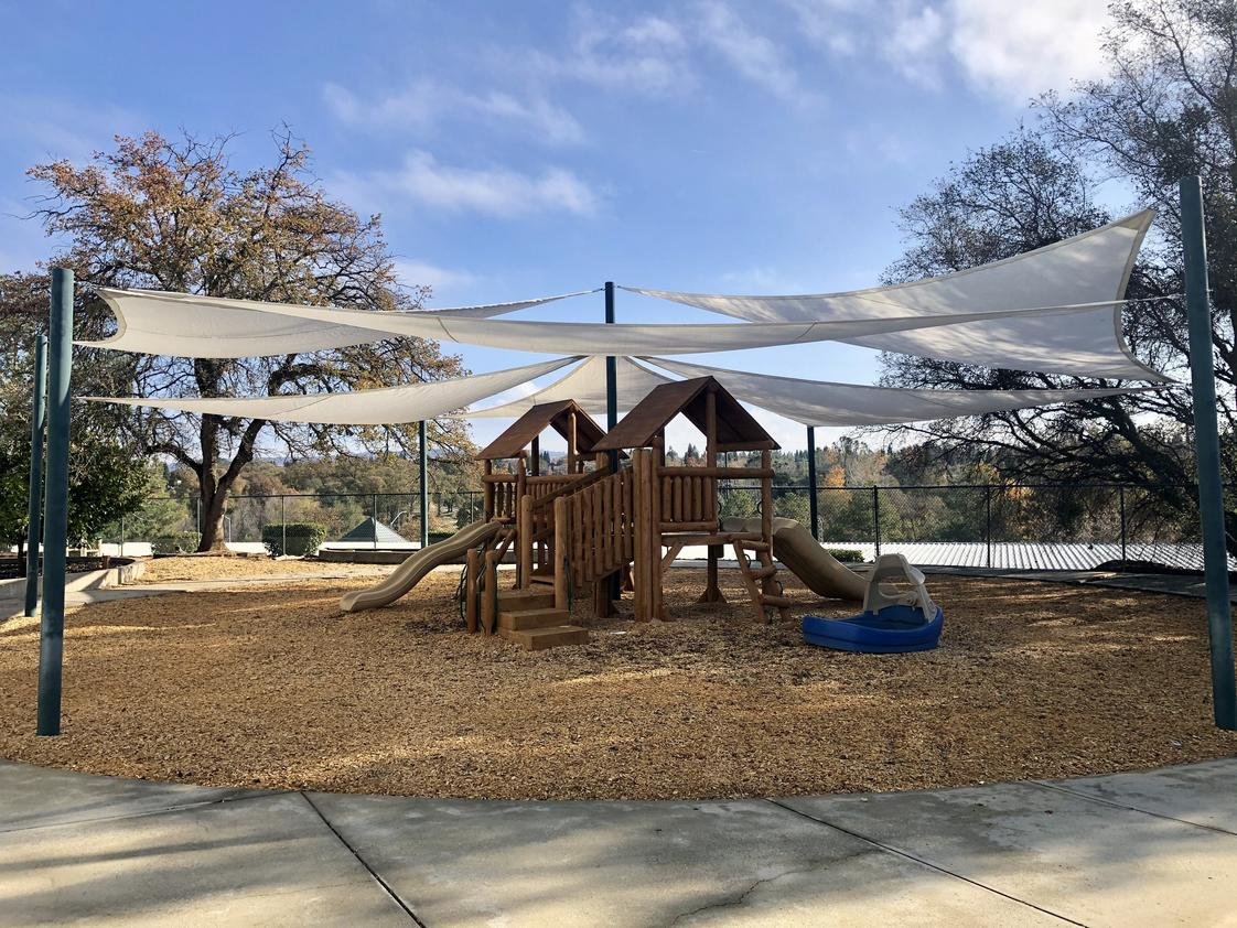 Empty playground with a wooden play structure, slides, and shade sails on a sunny day with trees in the background.