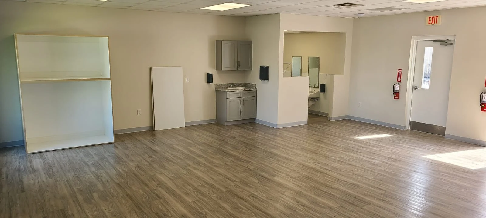 Empty room with wood-look flooring, corner shelving unit, small gray cabinet with granite countertop, and partial sink area with mirrors.