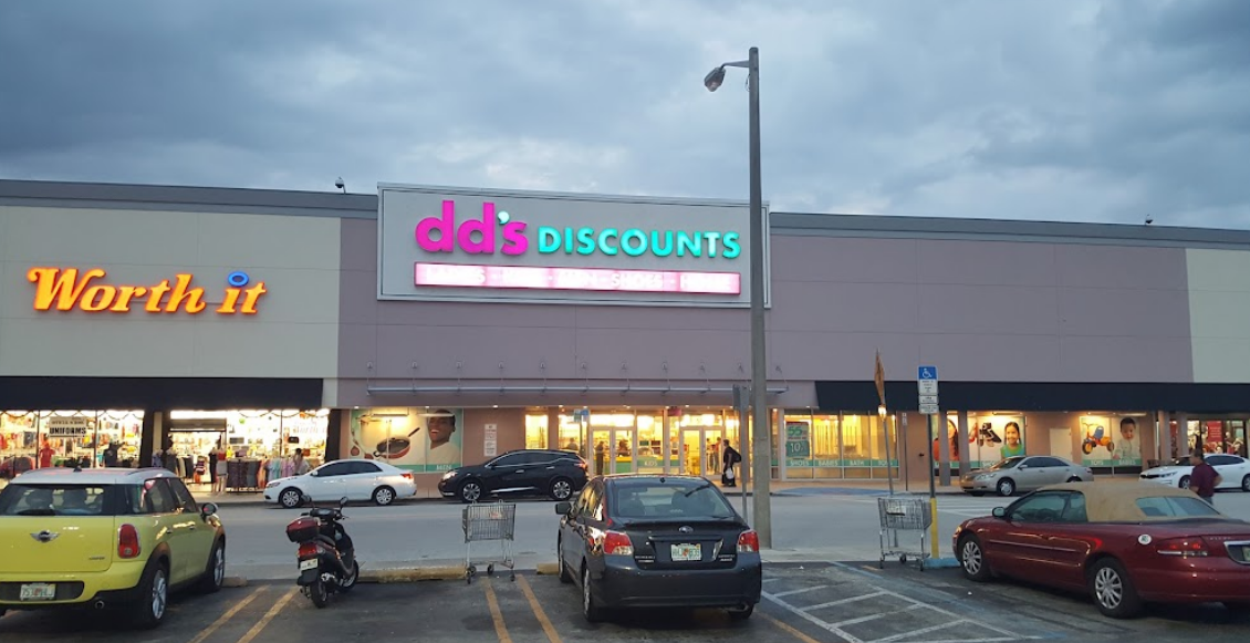 Exterior view of a shopping plaza featuring a sign for dd's Discounts store and a Worth it sign. The parking lot in front has several cars and a motorcycle, with shopping carts near the store entrance, under cloudy skies.