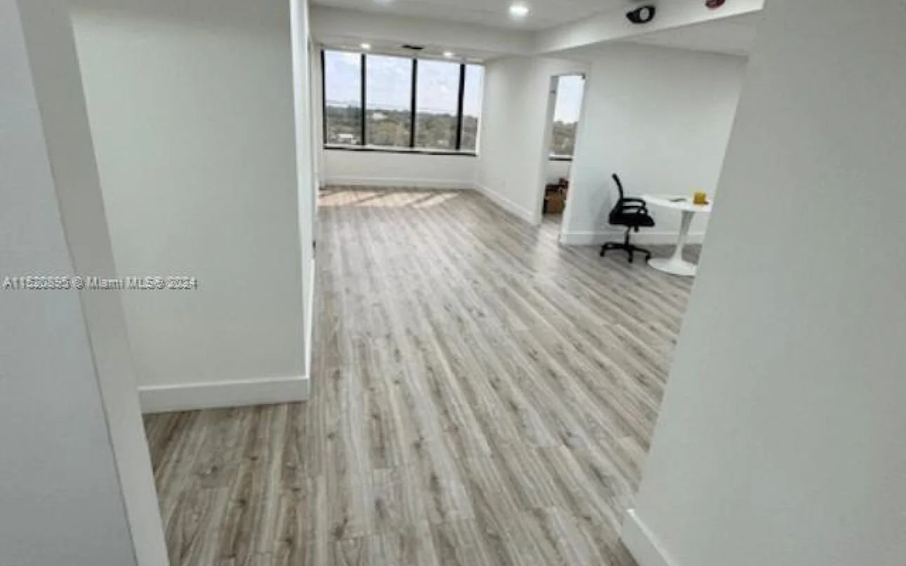 Empty modern apartment room with large window, light wood flooring, and a small white table with a black office chair.