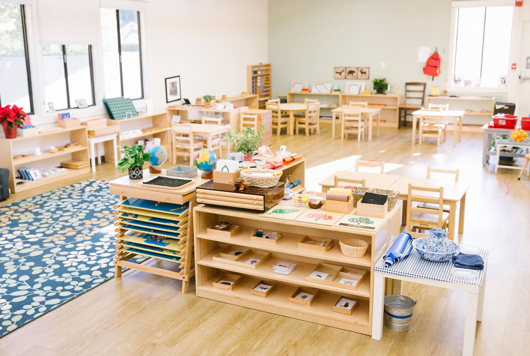Empty classroom with wooden tables and shelves, educational materials, and a decorative blue and white rug.
