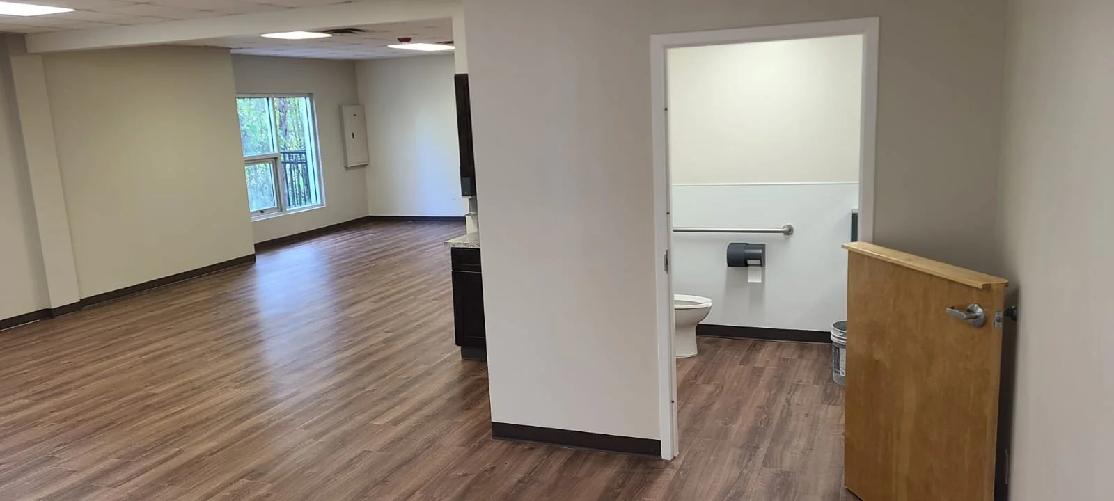 Empty room with wood-like flooring, white walls, and a small bathroom with accessible features visible through an open door.