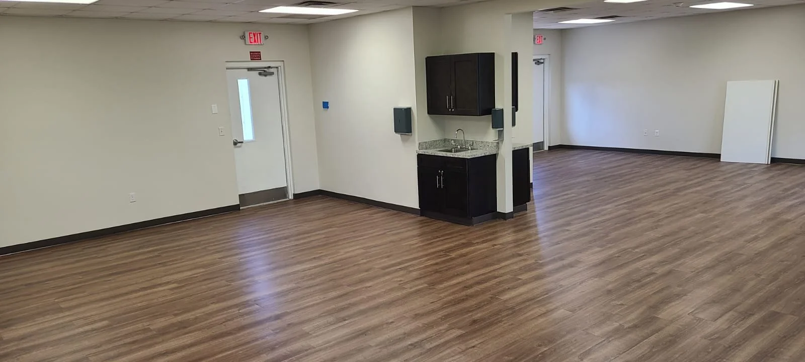 Empty room with wood flooring, a small kitchenette with dark cabinets and granite countertop, and a white wall with a door and exit signs.