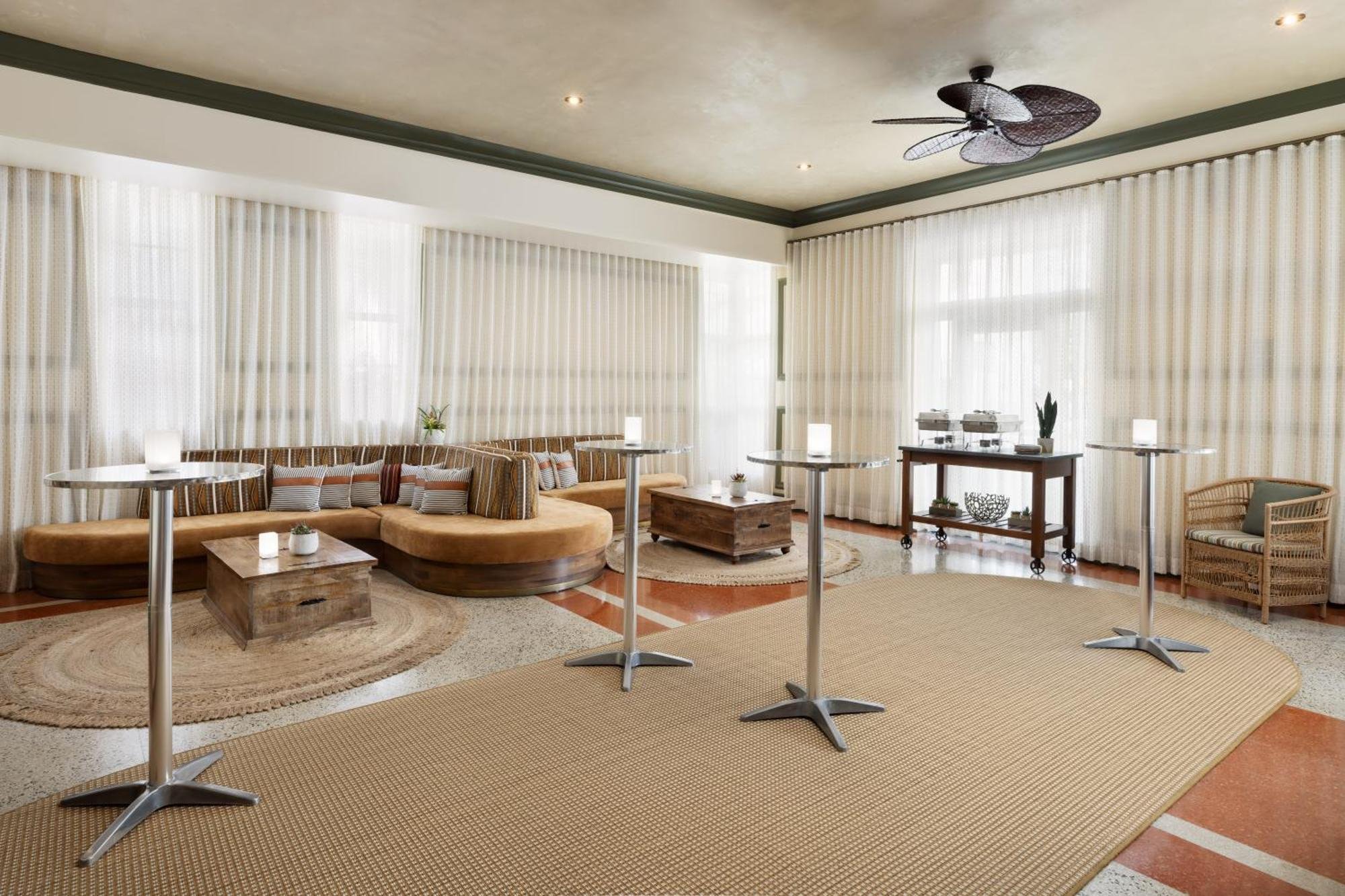 A spacious lounge area with beige curtains, a large cushioned sectional sofa with striped pillows, a wooden coffee table, four tall cocktail tables, a Rattan chair, and a buffet table with two chafing dishes. The room has recessed lighting and a ceil