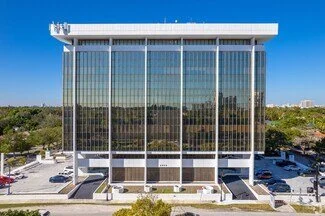A modern multi-story office building with a glass facade, surrounded by greenery and parking lots.