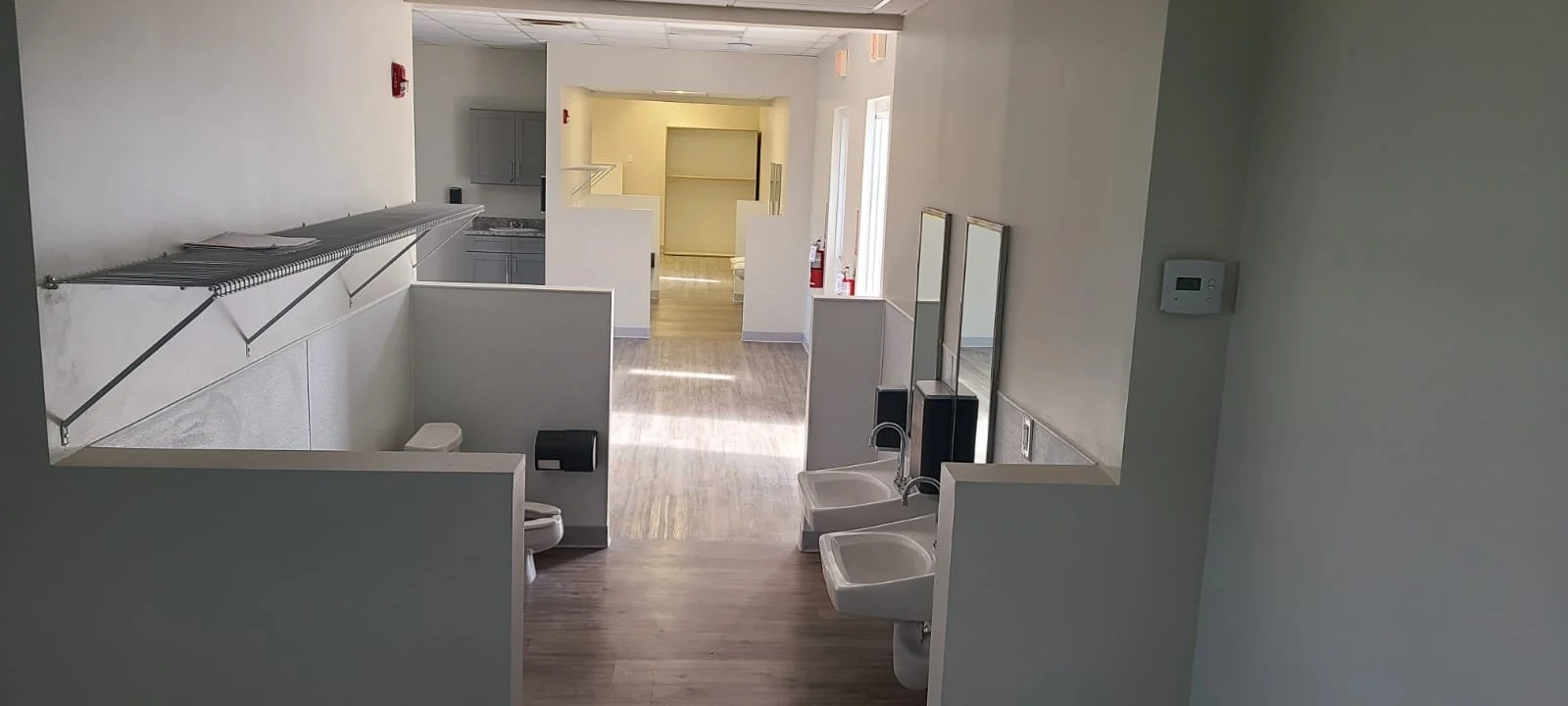 Empty room with accessible sinks, mirrors, a toilet, a trash can, and fire safety equipment, possibly a public restroom or facility.