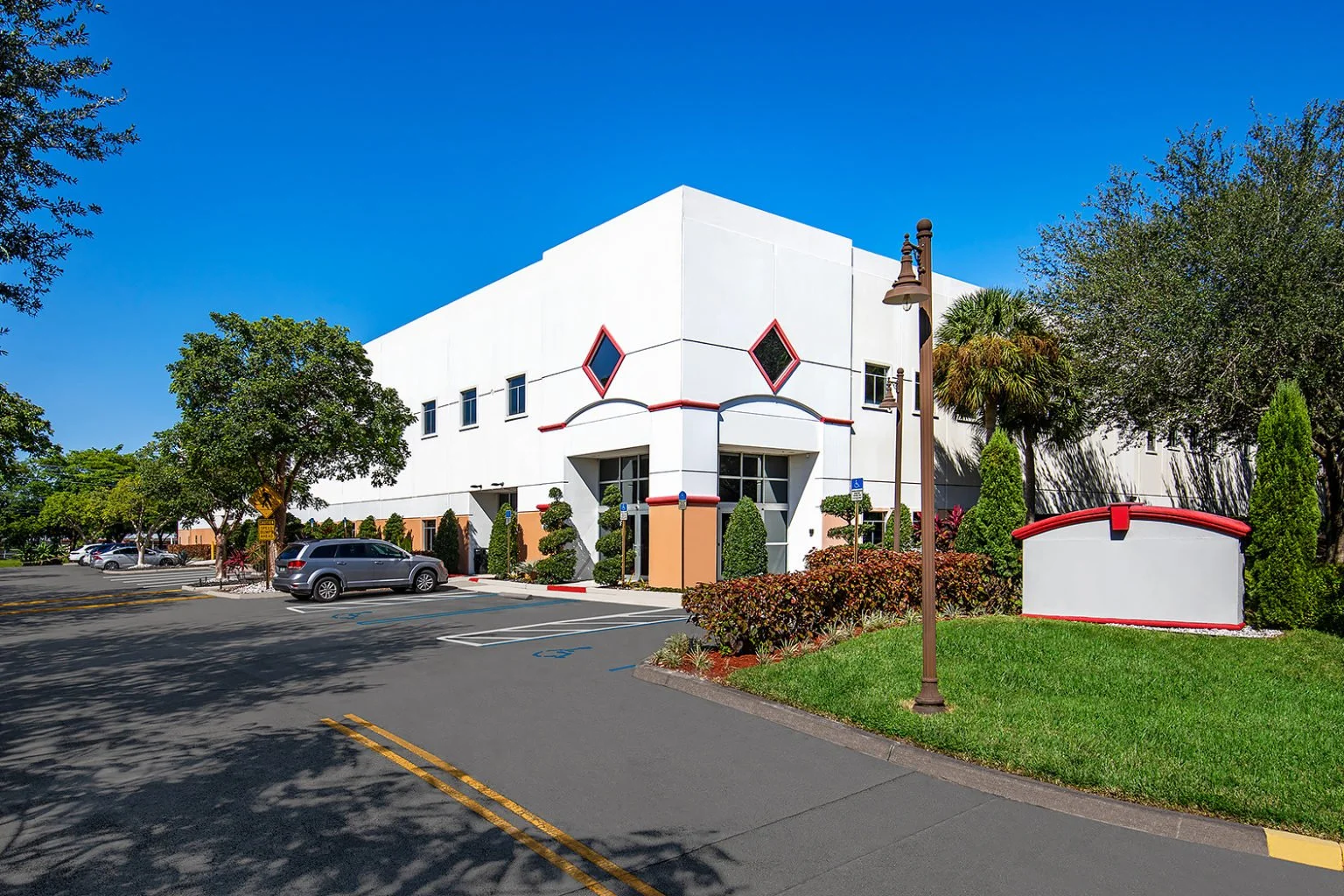 A modern white building with red and black accents, surrounded by trees and shrubbery, and with a parking lot in front.