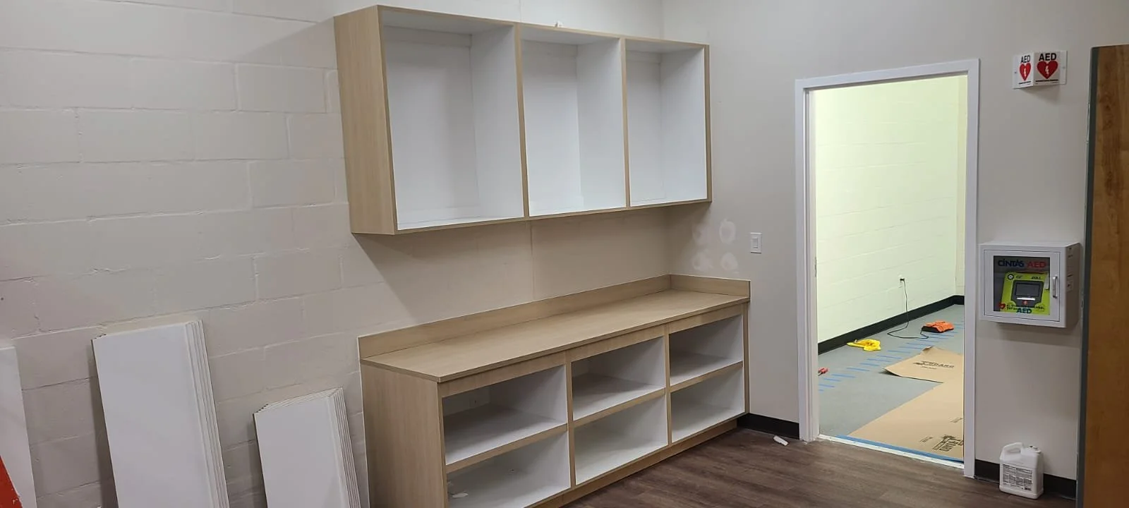 Empty wooden cabinet with open shelves in a room with a white brick wall and an open doorway leading to another room with tools and cardboard on the floor.
