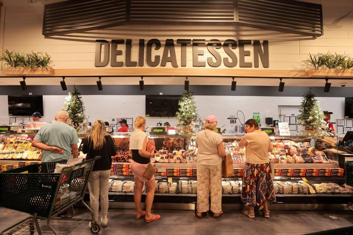 People shopping at a delicatessen counter with various meats and cheeses, inside a grocery store.