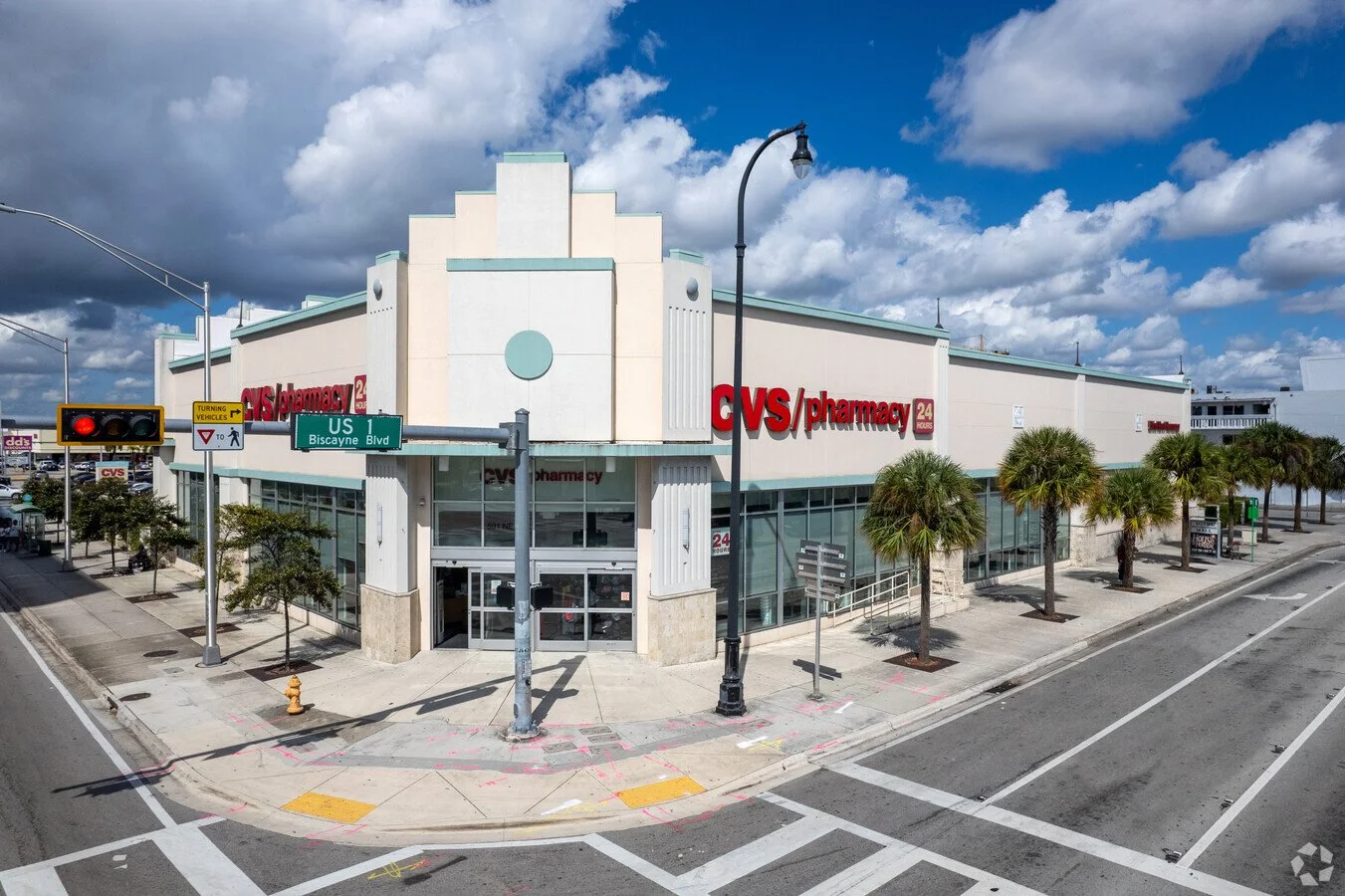 An exterior view of a CVS Pharmacy store at the corner of a street, showing the store's signage, palm trees lining the sidewalk, traffic lights, and a cloudy sky.