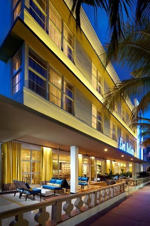 Exterior view of a hotel at dusk with yellow lighting, lounge chairs on the balcony, and palm trees nearby.