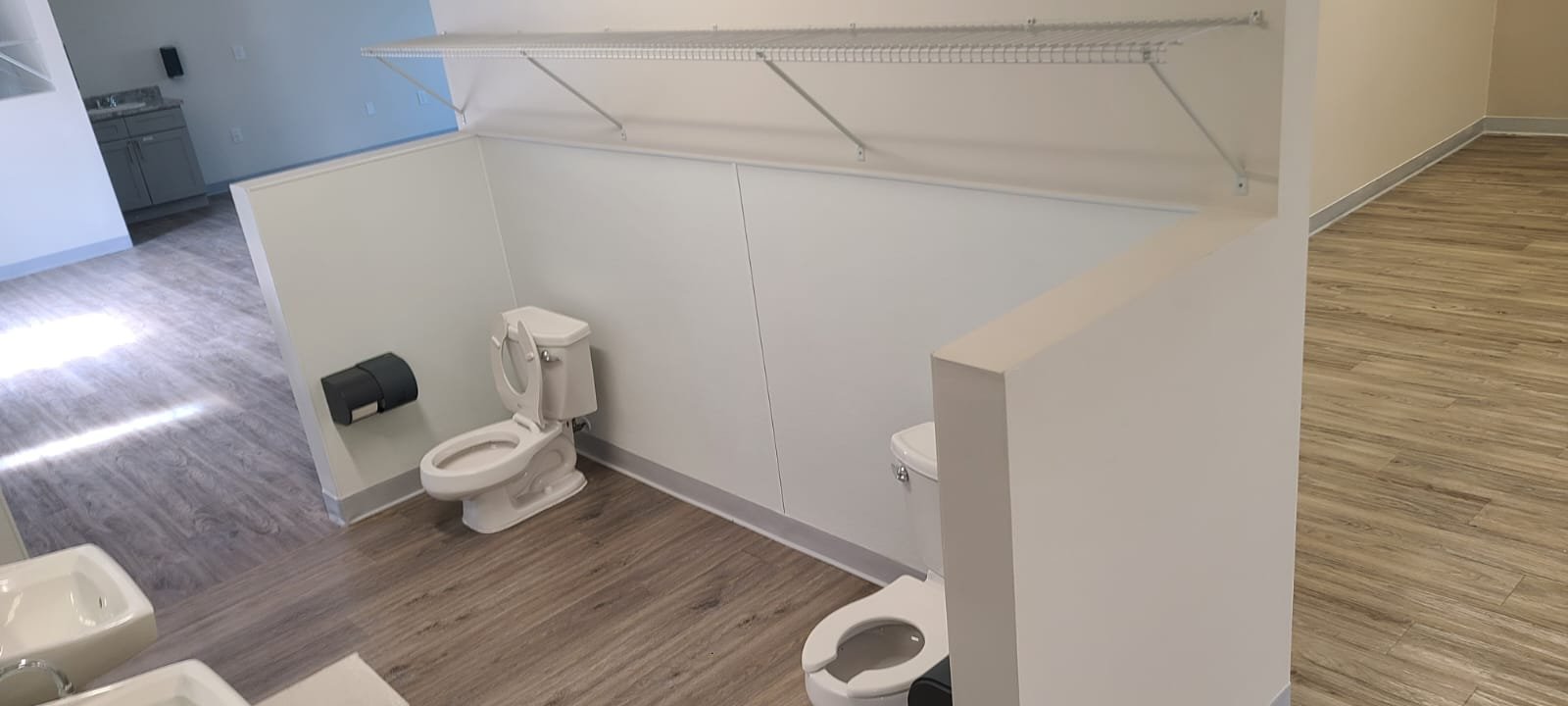 Public restroom with two toilets, a partial sink, and a high wall with a rack above the toilets.