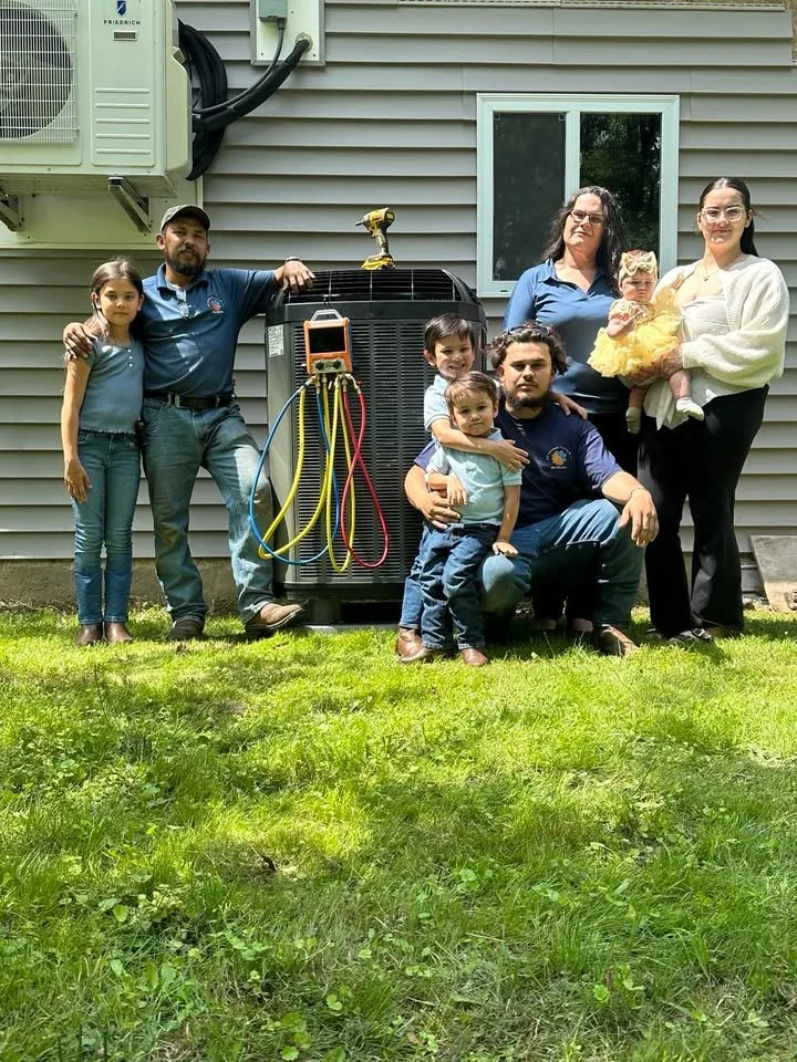 Group of seven people standing and sitting outside on grass in front of an air conditioning unit and house wall, including three children and four adults, with tools on the air conditioner.