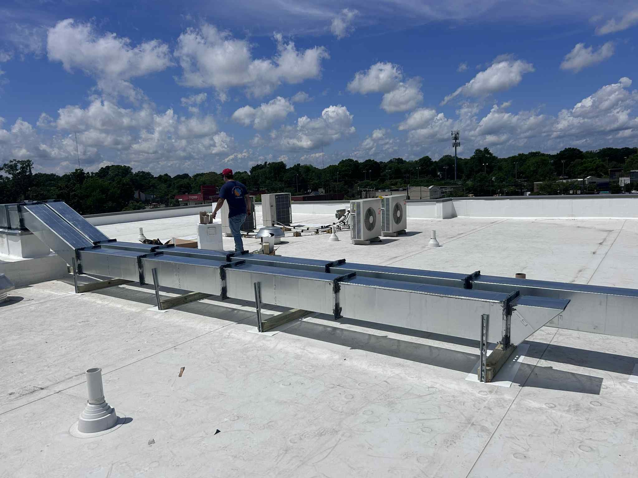 A construction worker on a rooftop with HVAC equipment and a skylight, under a partly cloudy sky.
