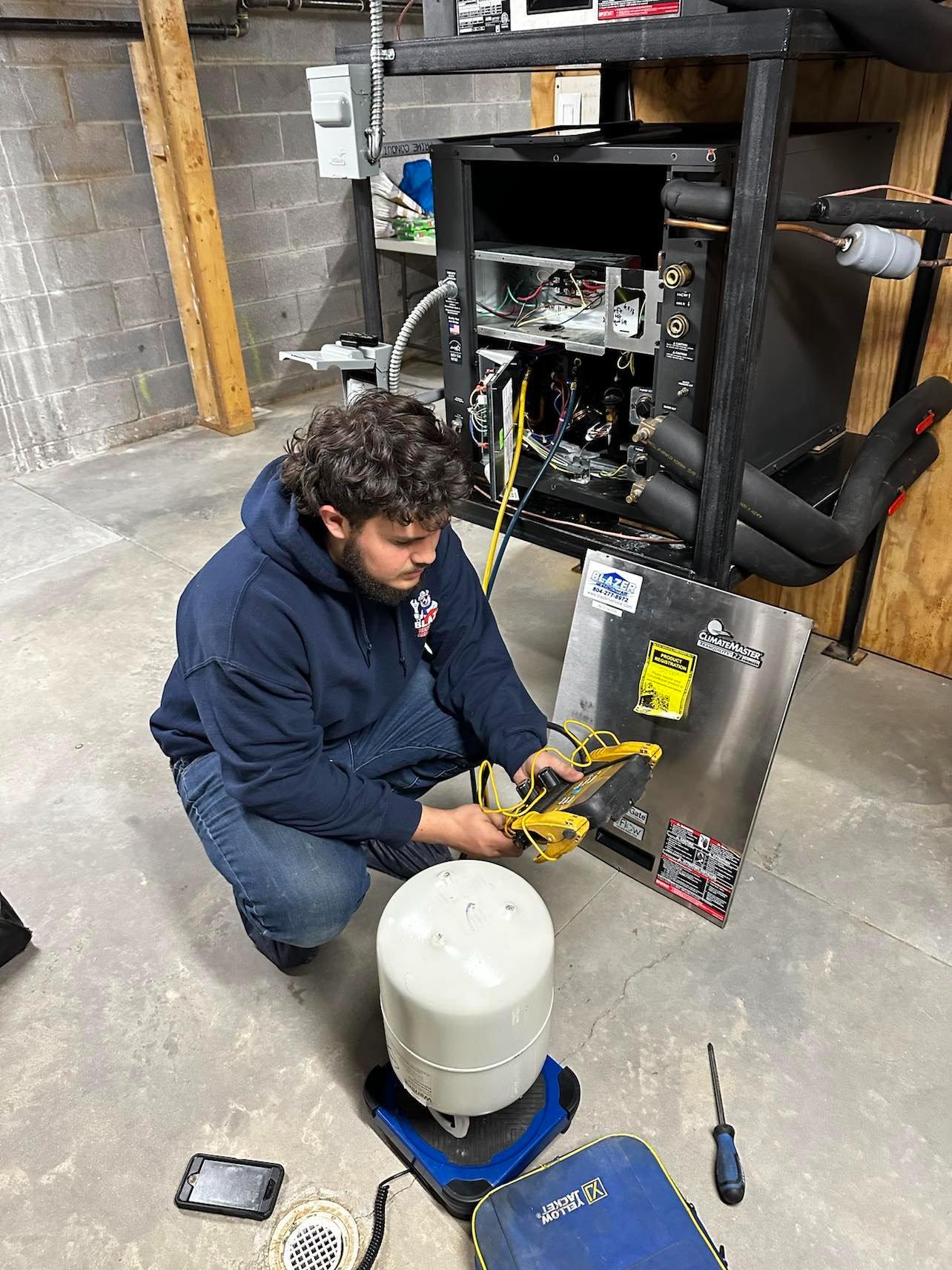 A technician working on an HVAC system or refrigeration unit, using a multimeter, inside a workshop or service area with concrete floors and brick walls.