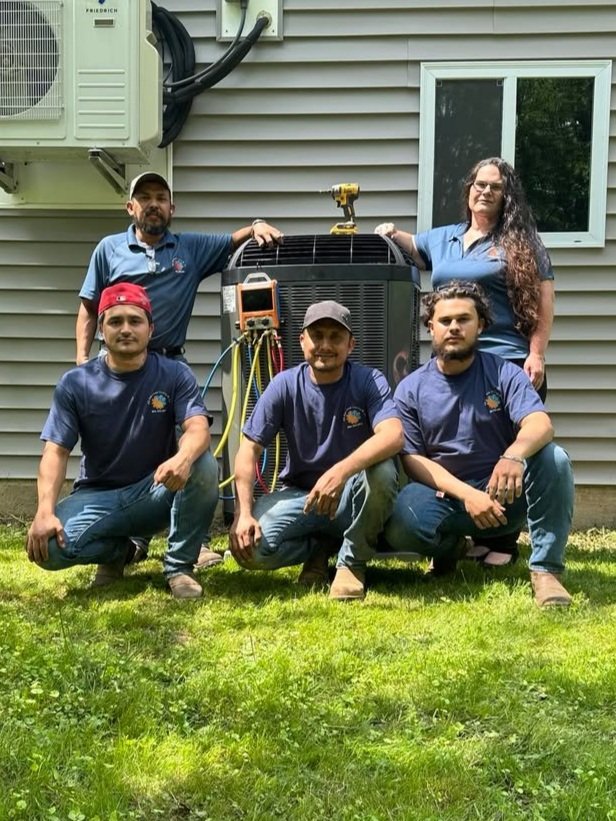 Five HVAC technicians in navy blue uniforms pose outside in front of an air conditioning unit, with two standing behind and three kneeling in front, on a grassy lawn beside a house with gray siding and a window.