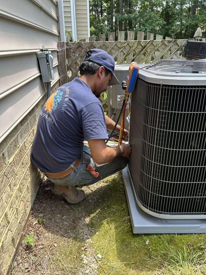 A technician working on an outdoor air conditioning unit beside a house, with a wooden fence and trees in the background.