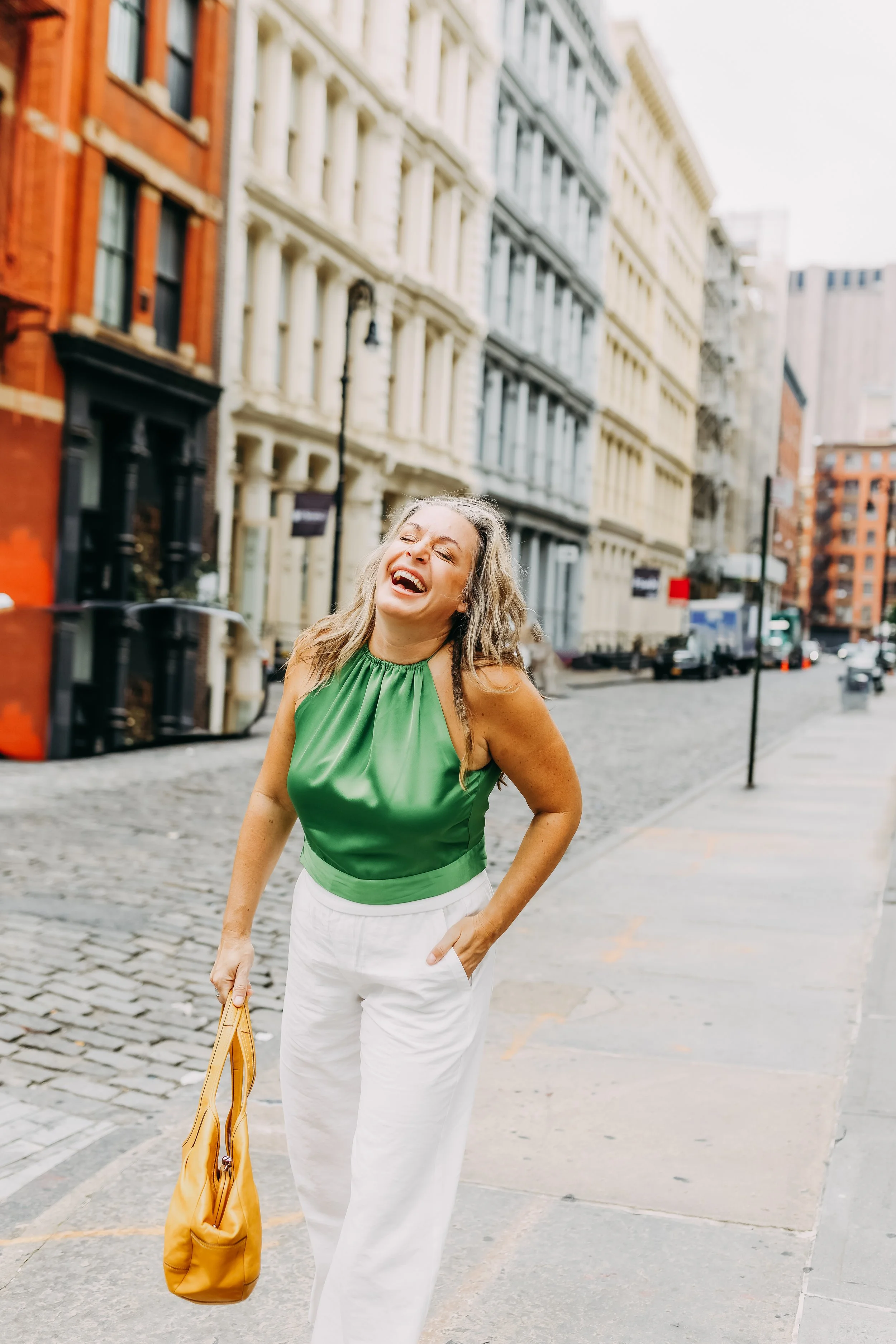 A woman with long gray hair, wearing a green sleeveless blouse and white pants, is standing on a city street, smiling and looking to her left, holding a yellow purse.