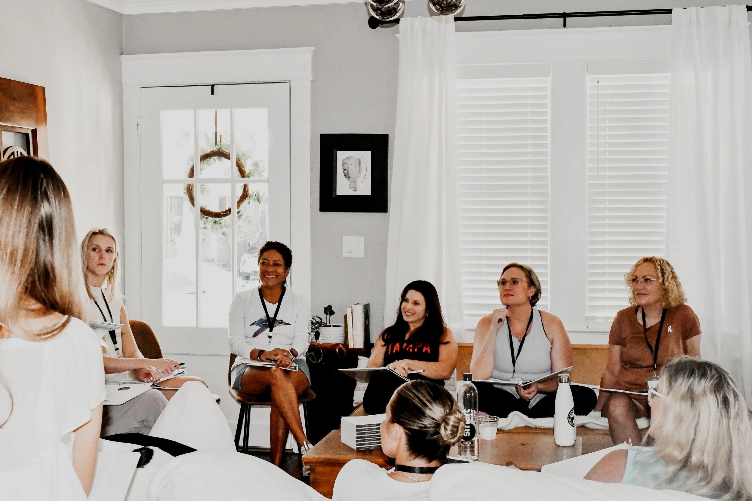 A group of women seated in a circle, engaged in a discussion or meeting inside a well-lit room with white walls and large windows with white blinds.