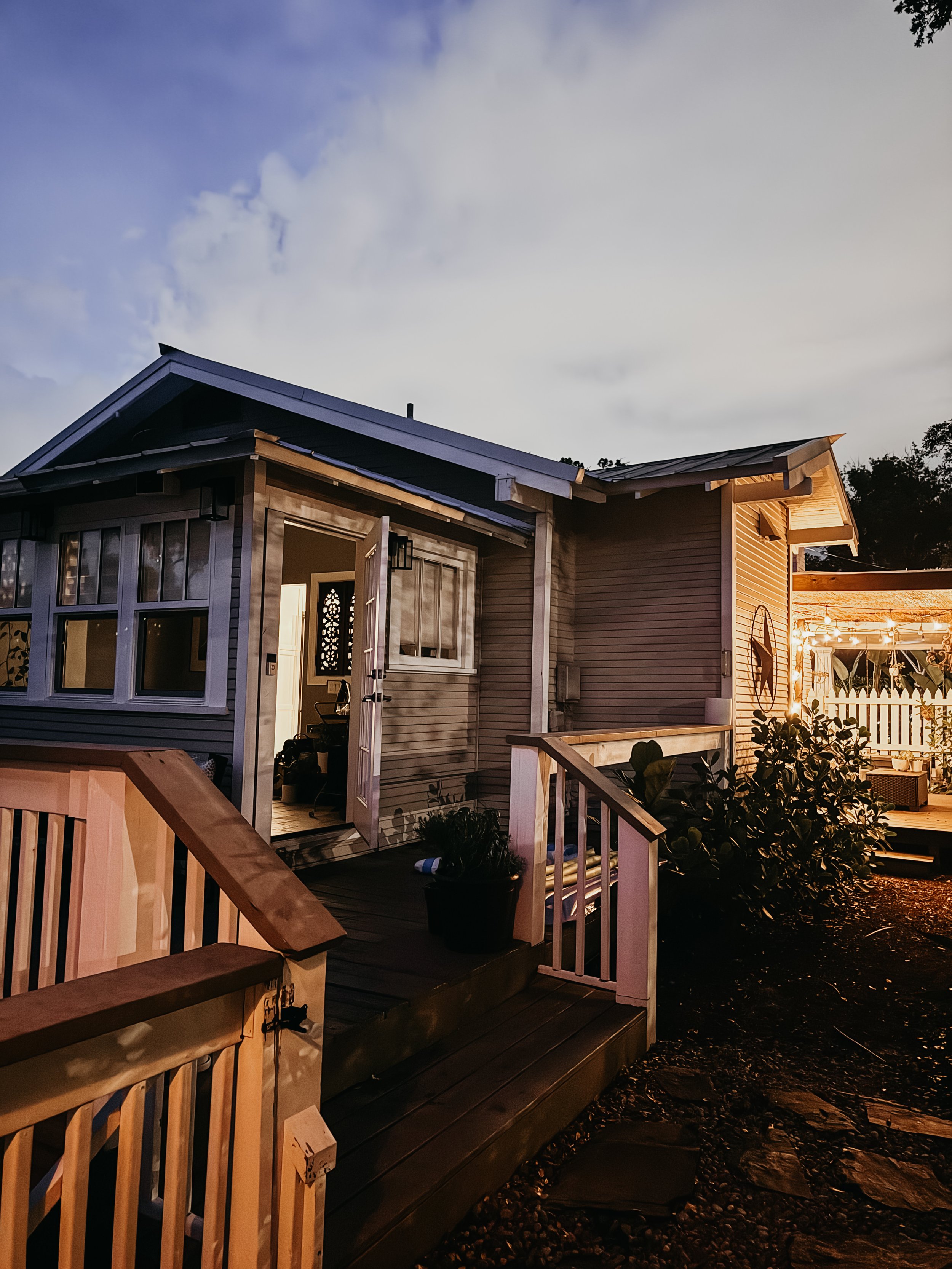Exterior view of a house with a porch, illuminated by warm lights, with a wooden railing, plants, and a sky at dusk.
