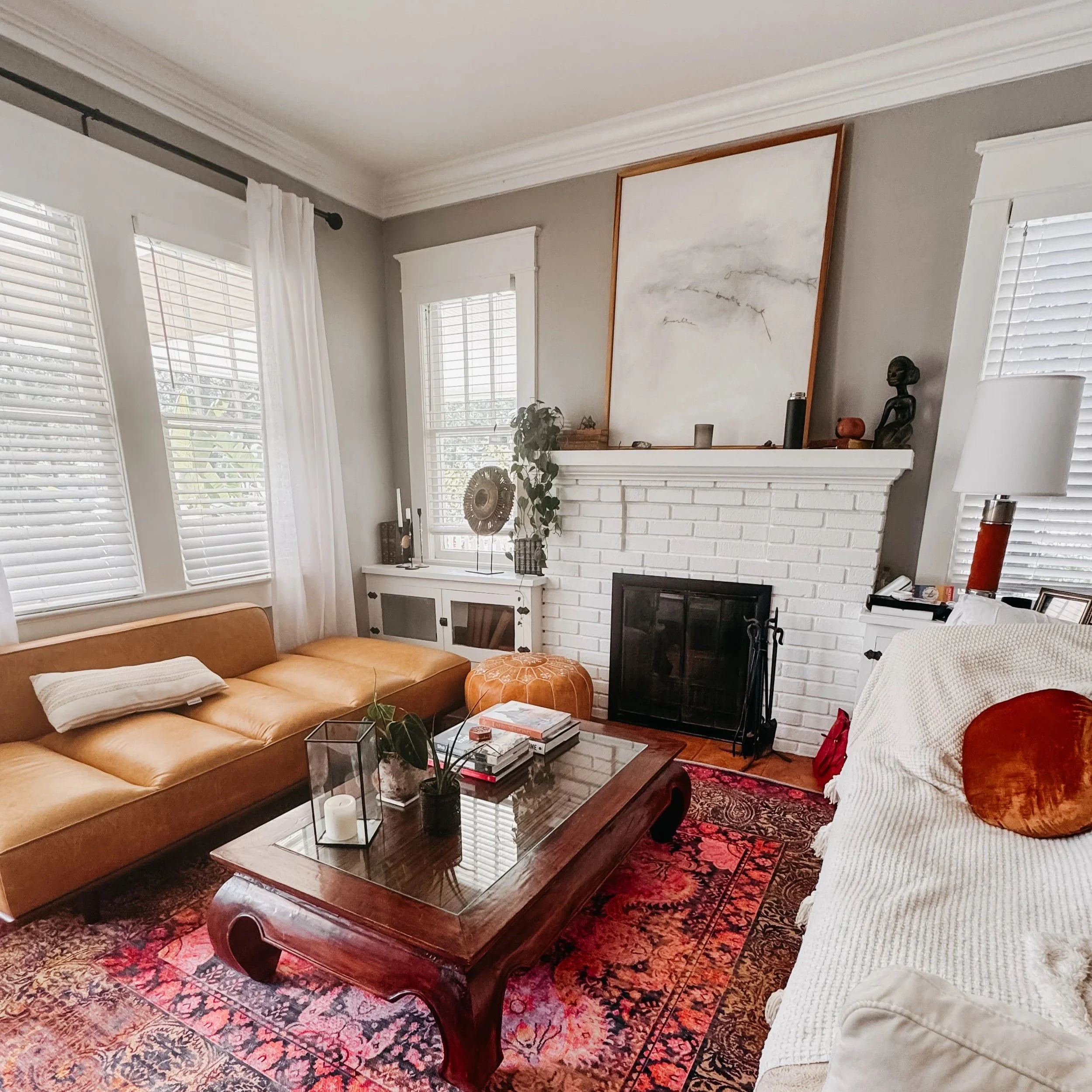 Living room with windows, white blinds, and a white brick fireplace decorated with artwork, sculptures, and plants. Contains a tan leather couch, a glass coffee table with books and a plant, and a patterned red and black rug.