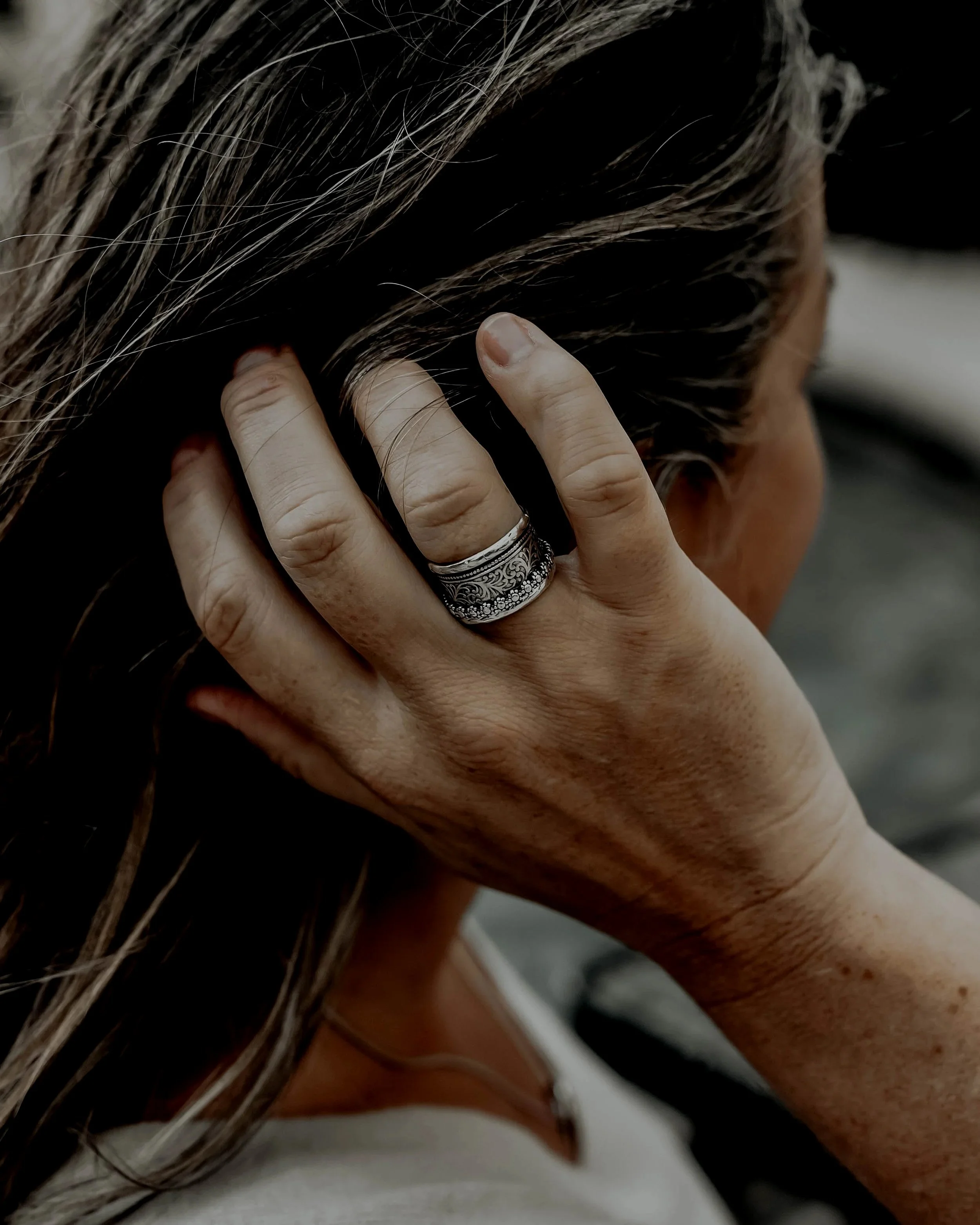A person with light to medium skin tone is holding their head with their hand, fingers resting on their scalp. The person has long hair with a mix of dark and gray strands. The hand is wearing a silver ring with intricate designs.